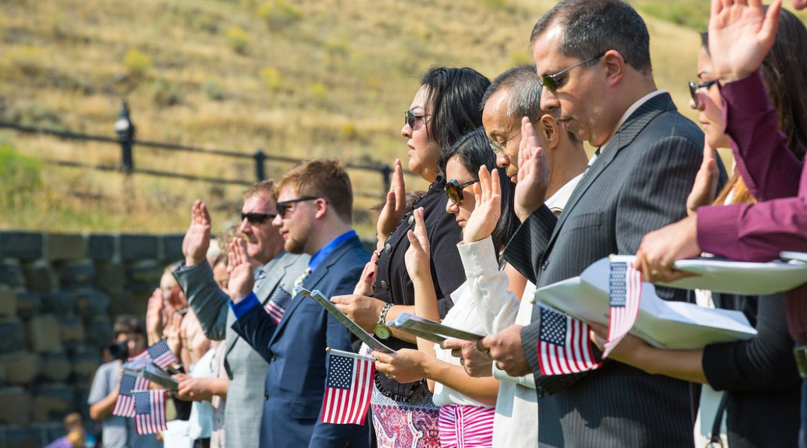 New American citizens holding small U.S. flags raise their right hands to take the Oath of Allegiance at an outdoor naturalization ceremony