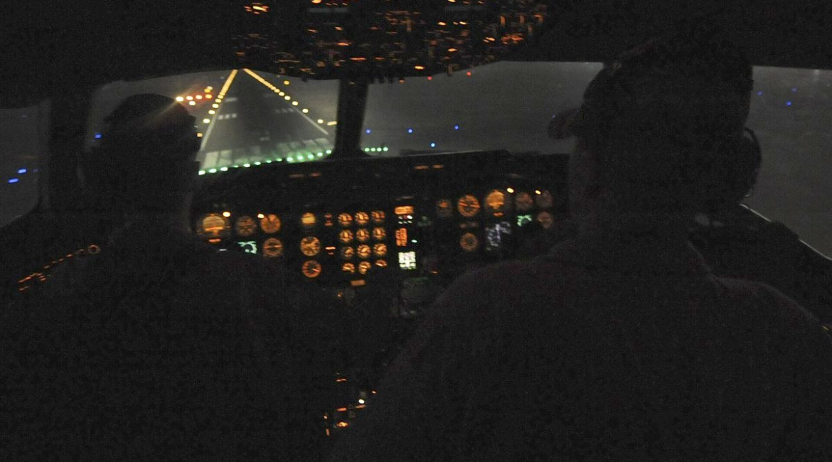 View from a darkened military aircraft cockpit at night, instrument panel glowing amber as the plane approaches a lit runway