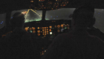 View from a darkened military aircraft cockpit at night, instrument panel glowing amber as the plane approaches a lit runway