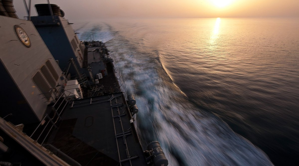 U.S. Navy destroyer cutting through calm water in the Strait of Hormuz at sunset, its wake trailing behind the ship