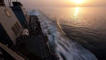 U.S. Navy destroyer cutting through calm water in the Strait of Hormuz at sunset, its wake trailing behind the ship