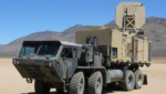Tan military truck carrying the Active Denial System's flat square antenna, parked on a desert flat beneath brown mountains and a clear blue sky