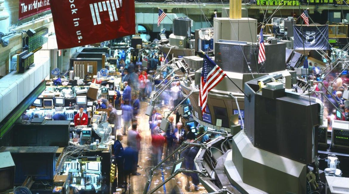 The busy trading floor of the New York Stock Exchange, with traders among banks of screens and an American flag hanging overhead